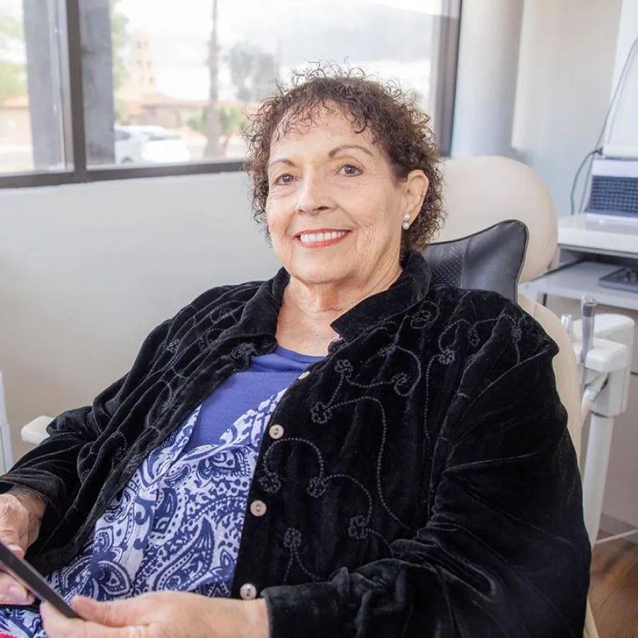 Smiling elderly woman sitting in a chair indoors wearing a black jacket and blue patterned shirt