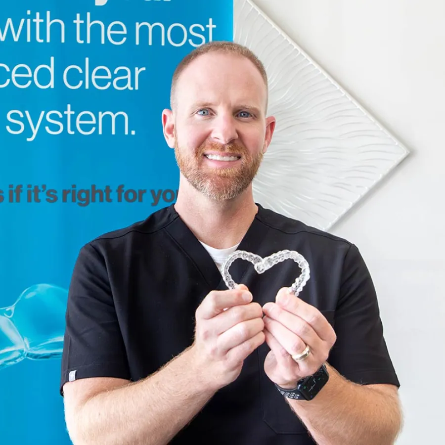 Smiling male dentist holding clear dental aligners shaped like a heart in a dental clinic.