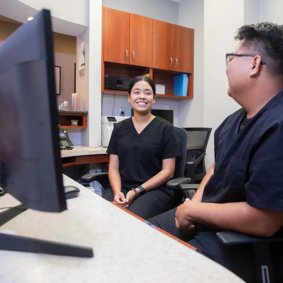 Two healthcare professionals in scrubs smiling and talking inside a medical office.