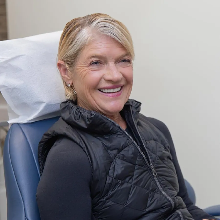 Smiling senior woman with short blonde hair sitting in a chair wearing a black vest indoors.