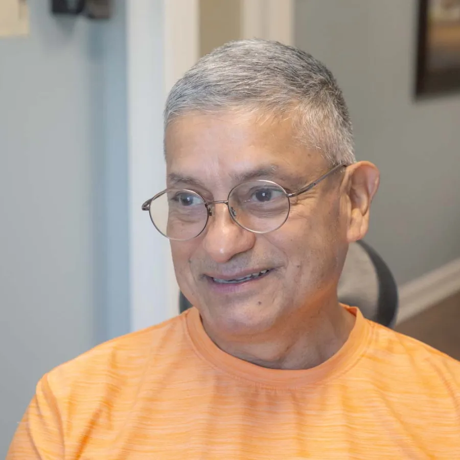 Smiling older man with gray hair and glasses wearing an orange shirt indoors in a home setting