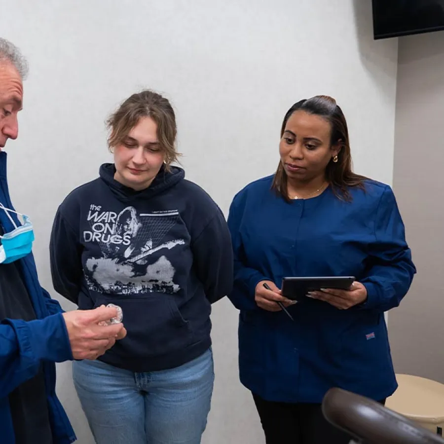 Three people engage in discussion indoors, one showing an object to two others, one holding a tablet.