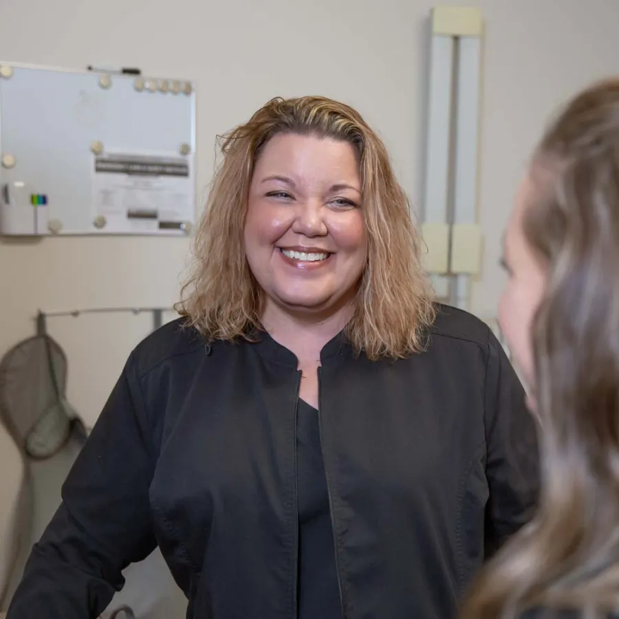 Smiling woman in black jacket talking to another person in indoor setting with medical or office equipment.