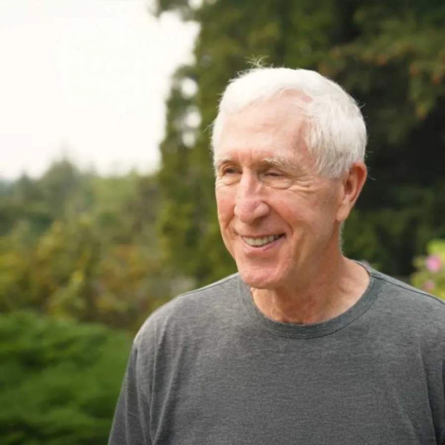 Elderly man with white hair smiling outdoors with blurred green foliage background