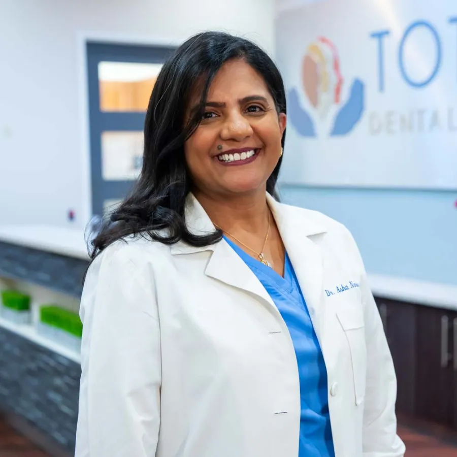 Smiling female dentist in white coat standing inside dental clinic lobby with Total Dental Care sign behind her.