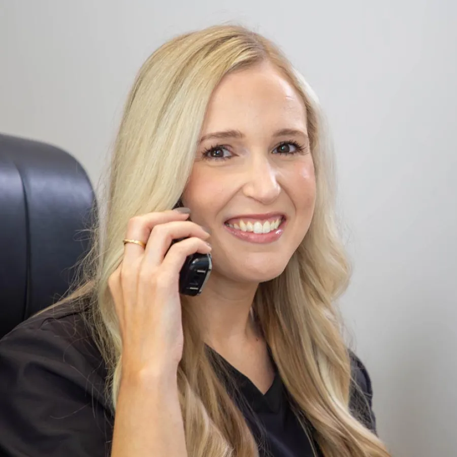 Smiling blonde woman in black top holding a phone to her ear against a neutral background.