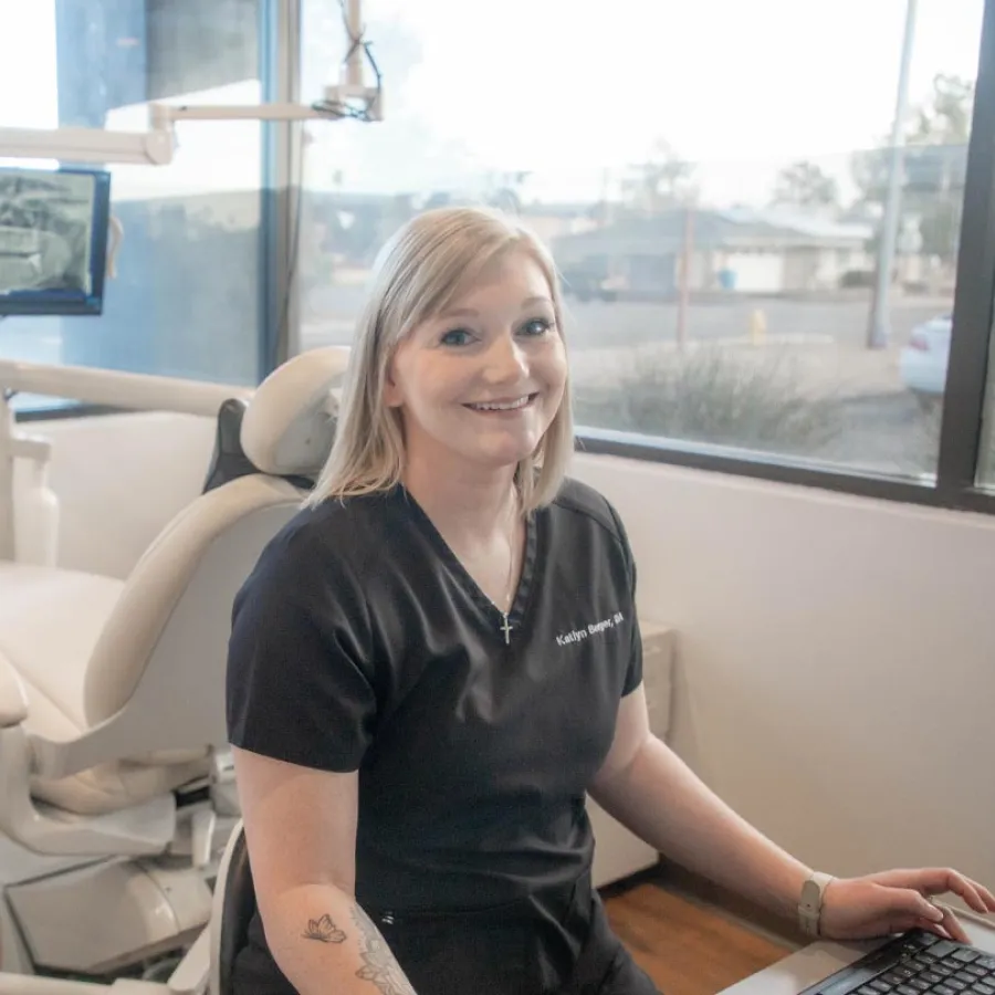 Smiling female dental professional sitting at a desk in a bright clinic with dental chair in background