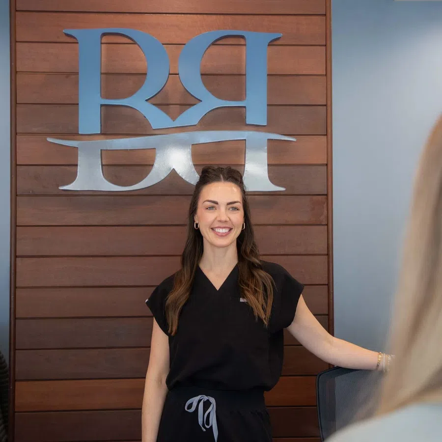 Smiling woman in black medical scrubs stands against a wooden wall with a large stylized logo behind her.