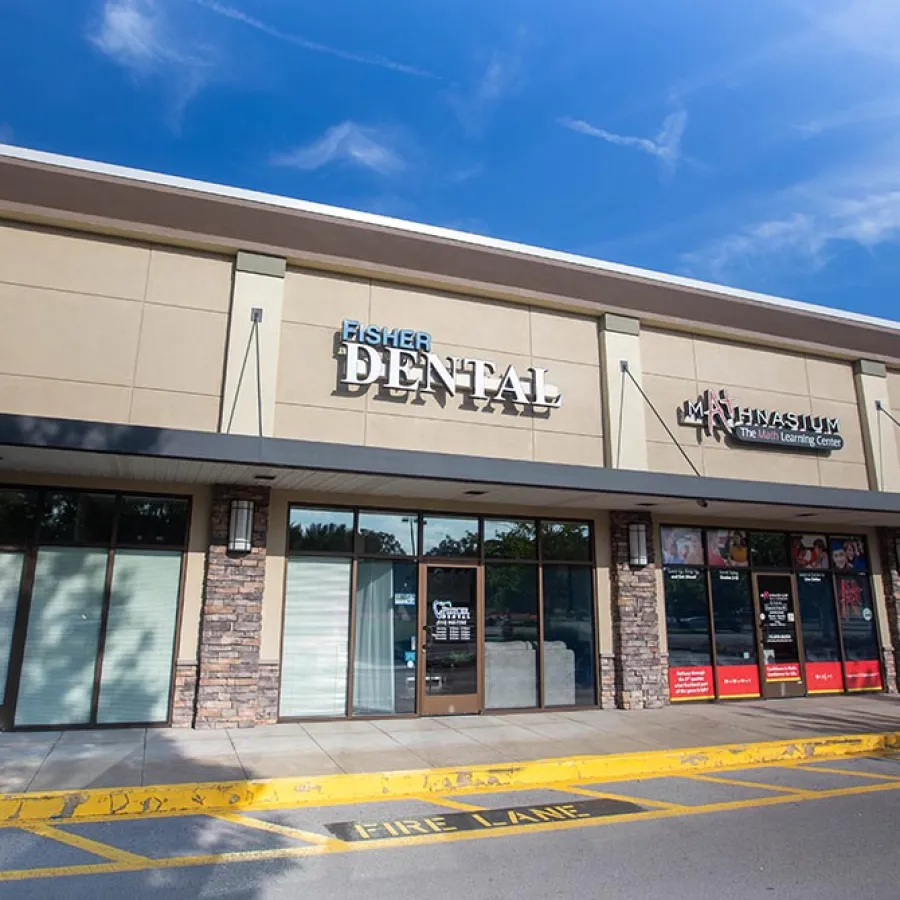 Exterior view of a shopping strip featuring Fisher Dental, a learning center, and a Thai restaurant under a clear blue sky.