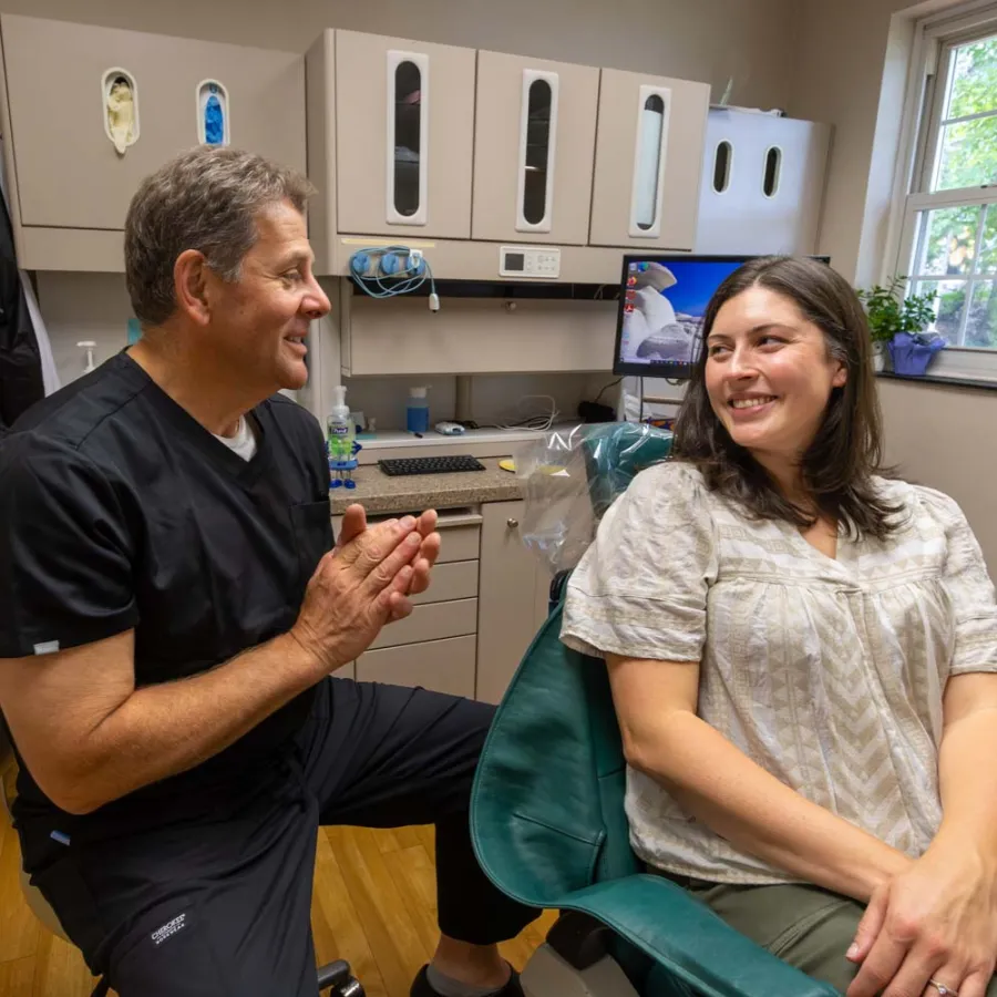 Dentist in black scrubs talking with female patient smiling in dental office with equipment and window light.