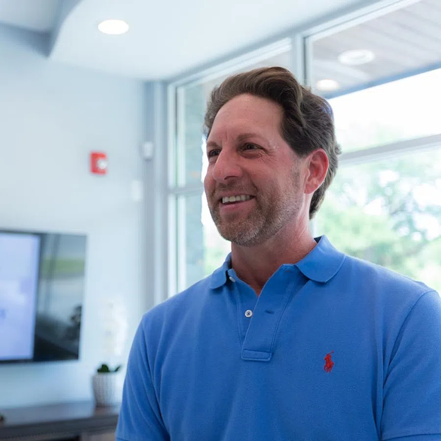 Smiling man in a blue polo shirt standing indoors near large windows with natural light.