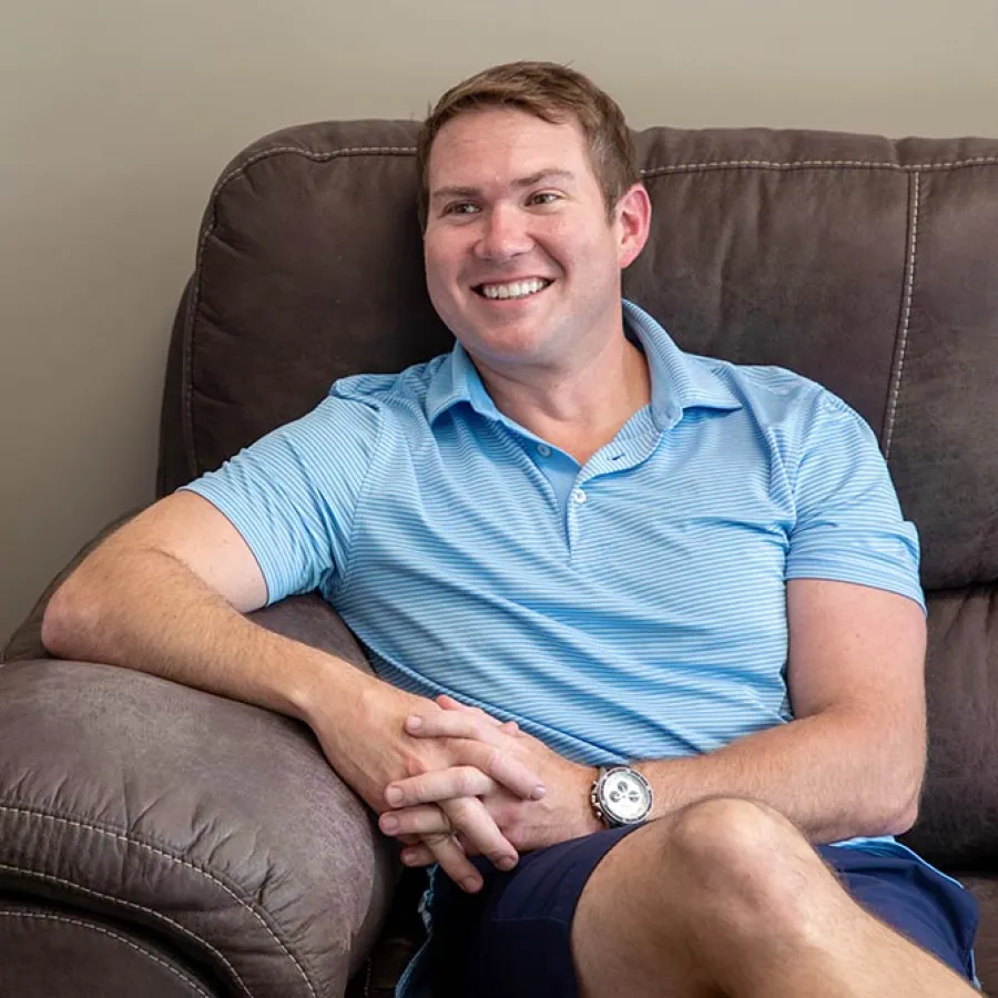 Smiling man in a blue polo shirt sitting relaxed on a brown couch with a fan on a side table nearby.