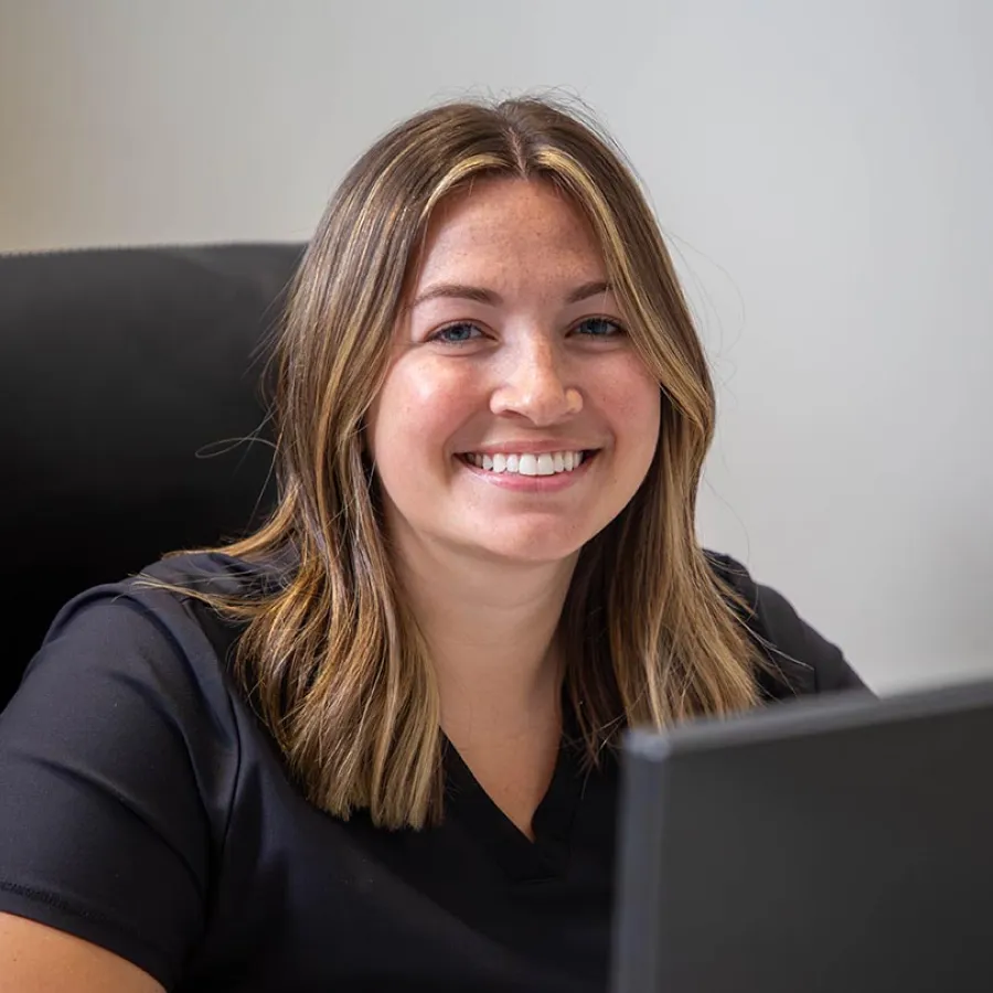 Smiling young woman with shoulder-length hair sitting at a desk in front of a laptop in an office setting.