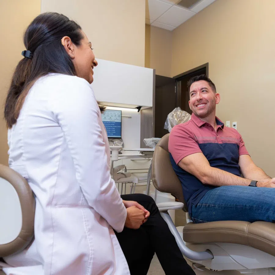 Male patient sitting in dental chair smiling and talking with female dentist in clinic room.