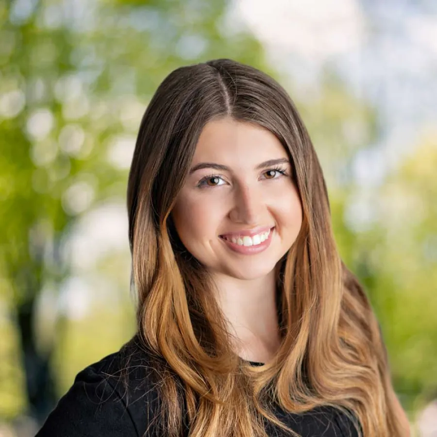 Young woman with long brown hair smiling outdoors with blurred green background wearing a black shirt