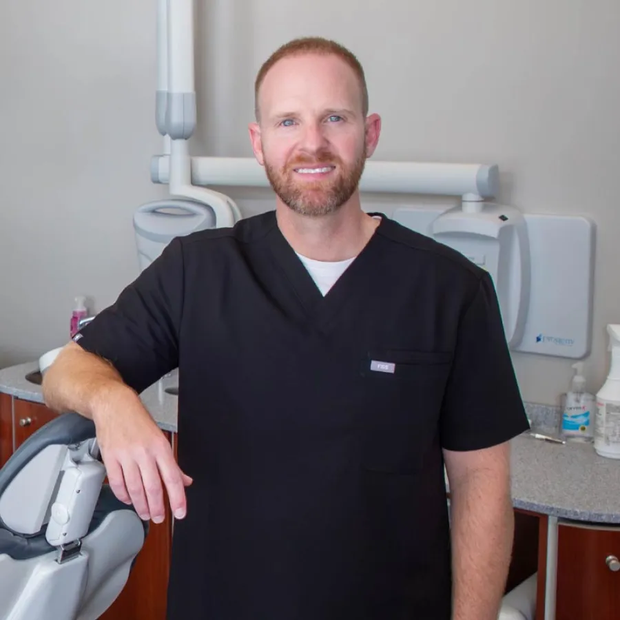 Male dentist in black scrubs smiling in dental office next to dental chair and equipment