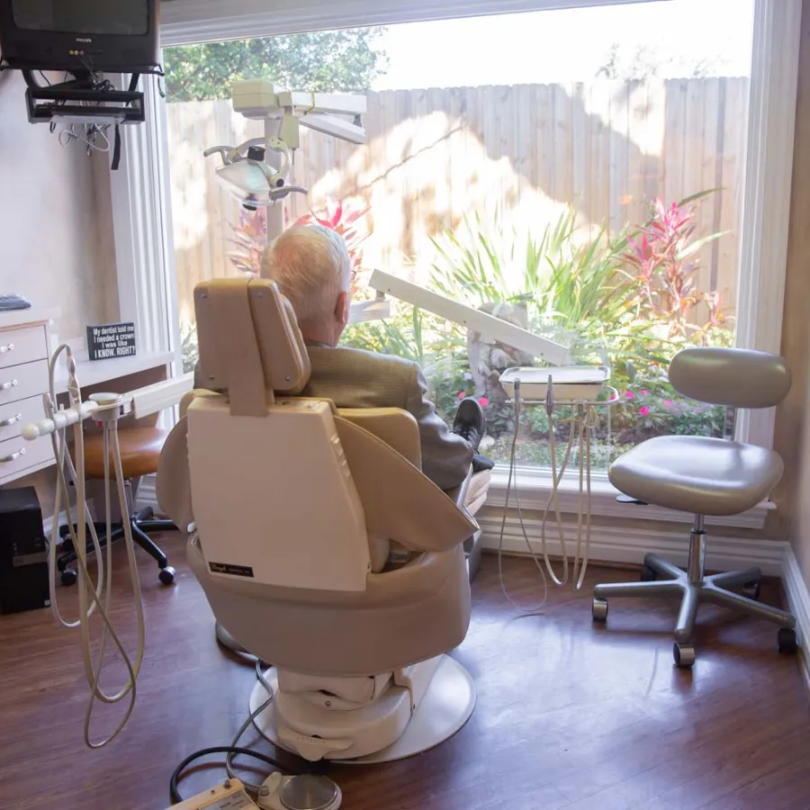 Senior man seated in a dental chair facing a large window with garden view in a modern dental office.