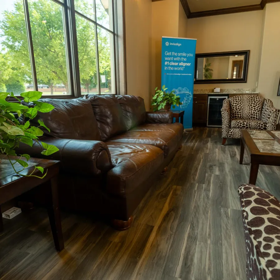 Comfortable waiting room with leather sofa, patterned chairs, plants, large windows, and Invisalign promotional banner.