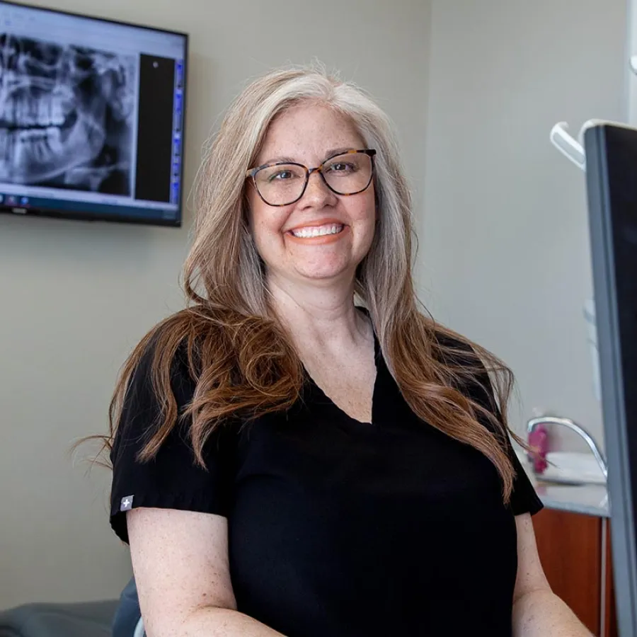 Smiling female dentist with glasses in black scrubs sitting in dental office with dental X-ray on screen behind her