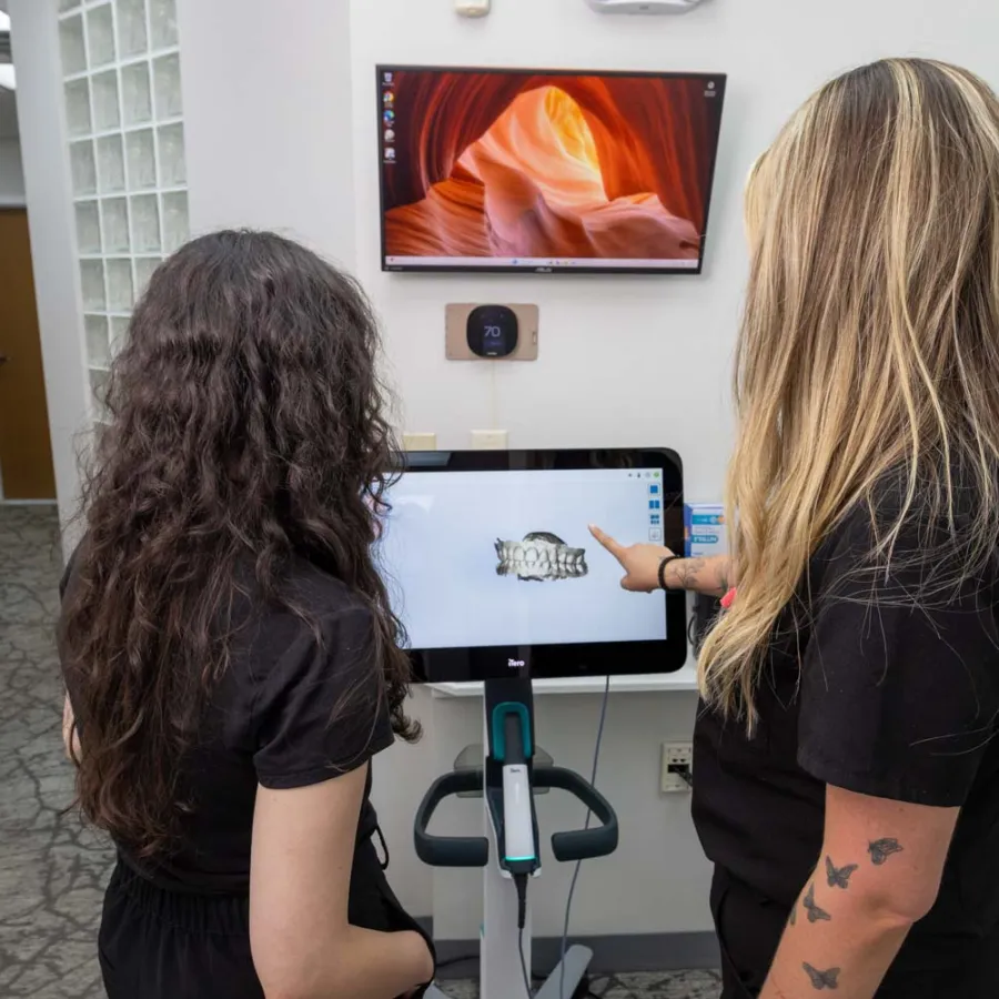 Two women view a dental 3D scan on a screen in a modern office with a wall-mounted monitor and thermostat.