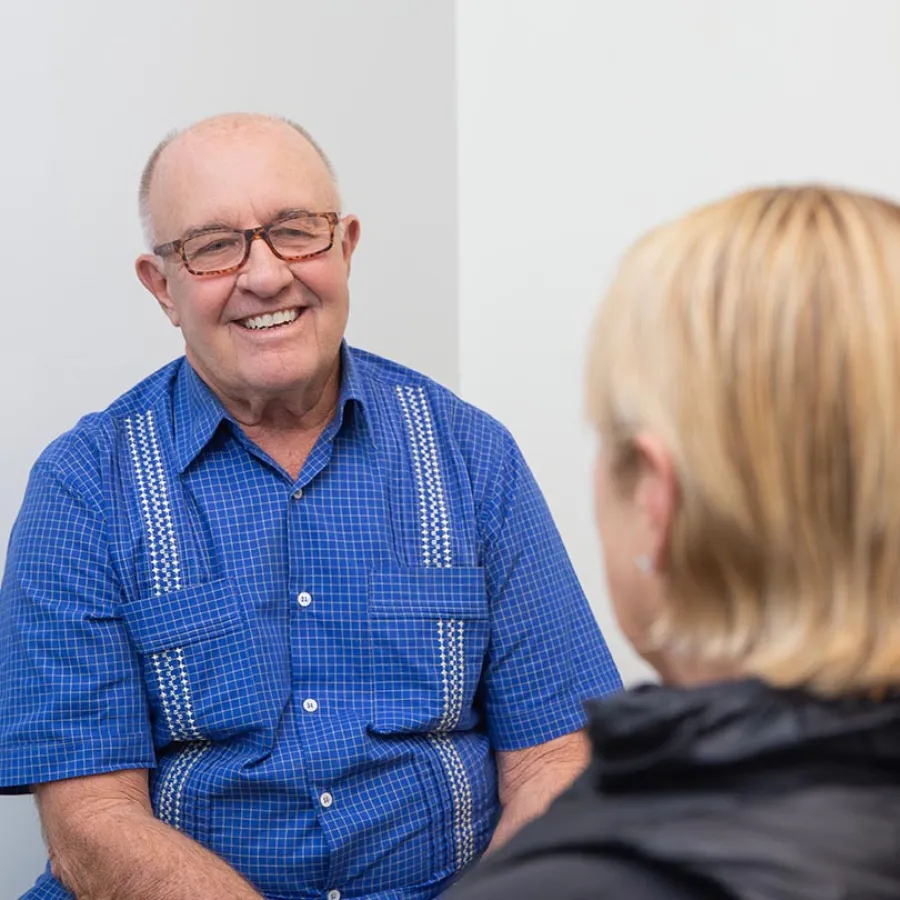 Smiling elderly man in blue checkered shirt talking to a blonde woman in a casual indoor setting.