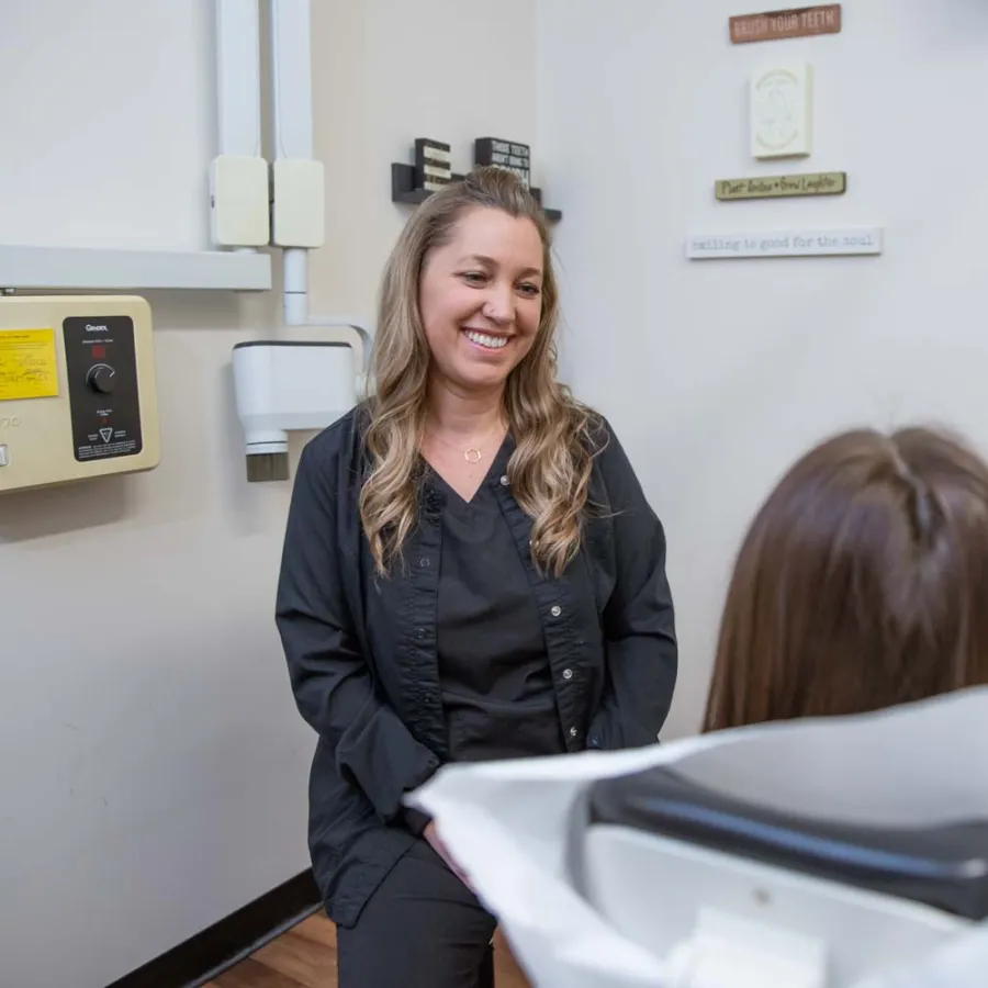 Dental hygienist smiling and talking to patient seated in dental chair in a clean office room.