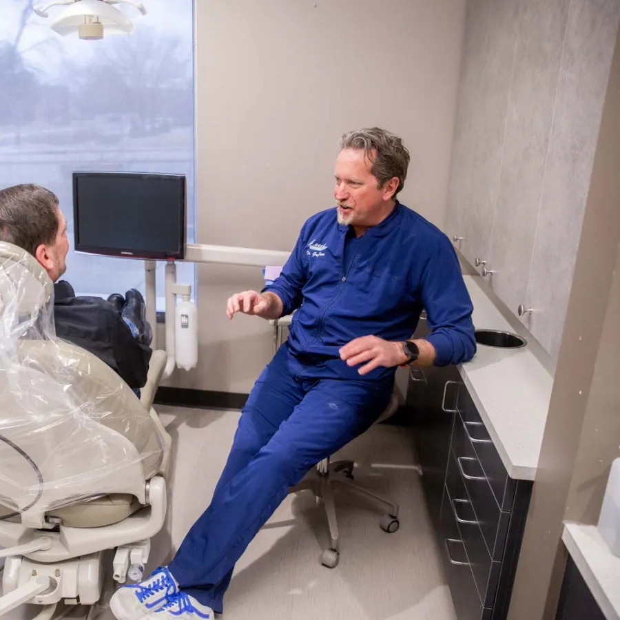 Dentist in blue scrubs consulting a male patient seated in a dental chair in a modern clinic room.