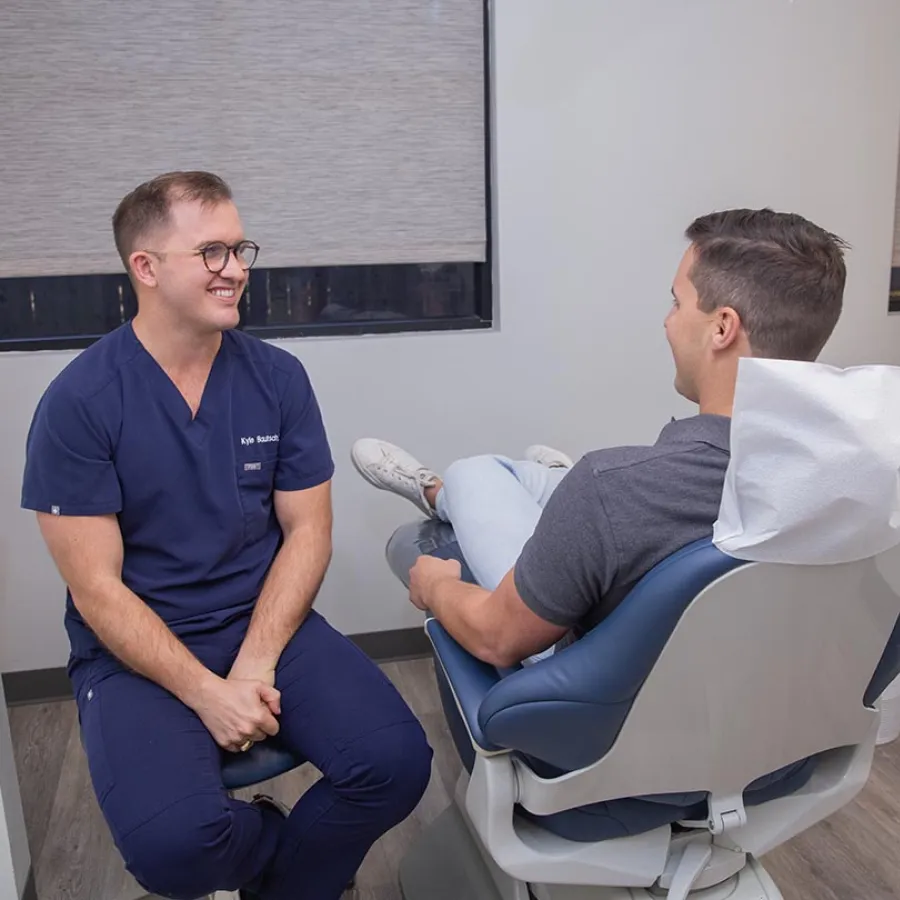 A dentist in navy scrubs smiling and talking to a male patient seated in a dental chair during consultation.