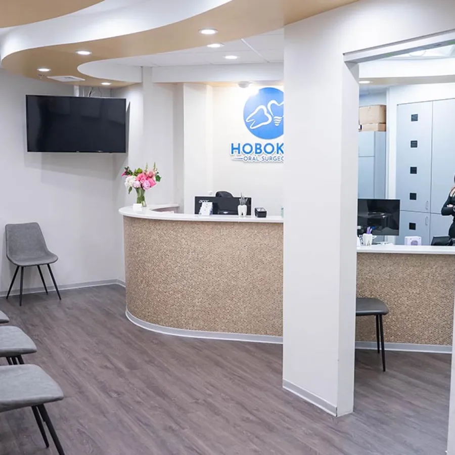 Modern dental clinic waiting area with gray chairs, wooden floor, curved reception desk, and wall-mounted TV.