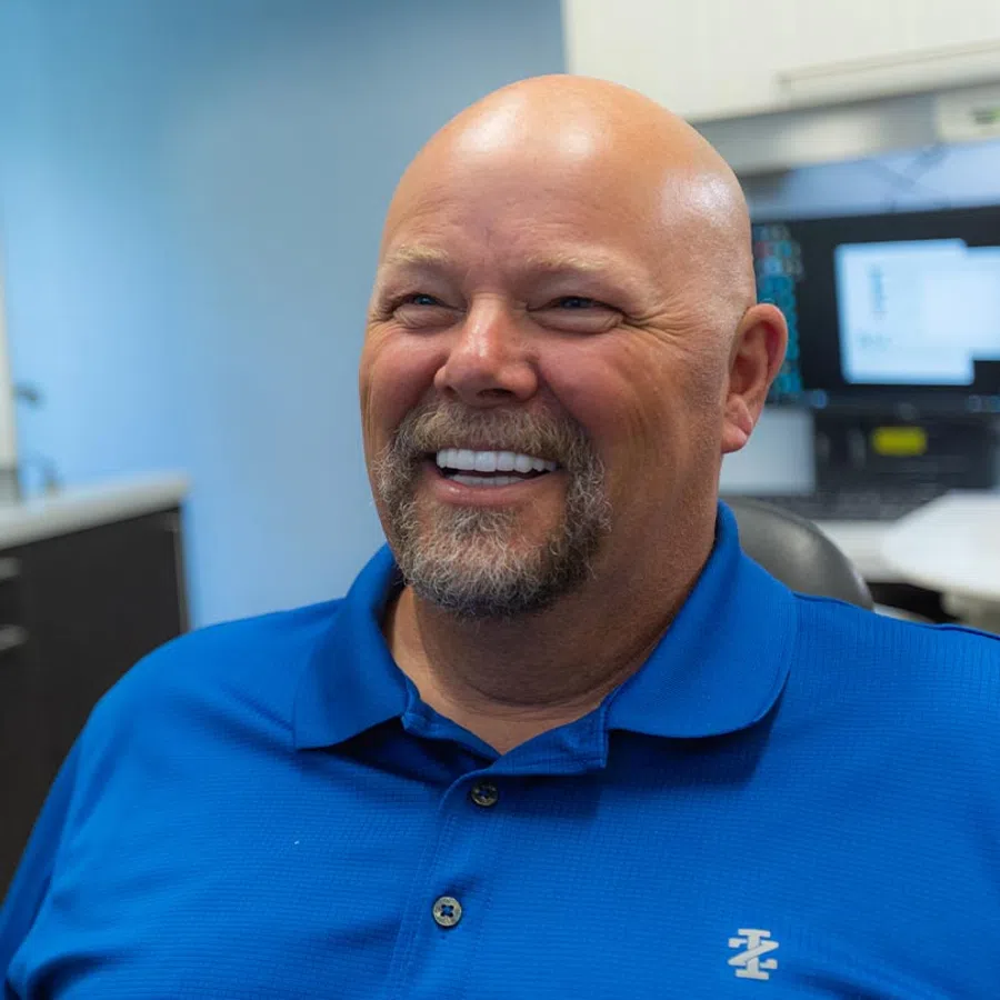 Smiling bald man with beard wearing a blue polo shirt sitting in an office with computer screens in the background
