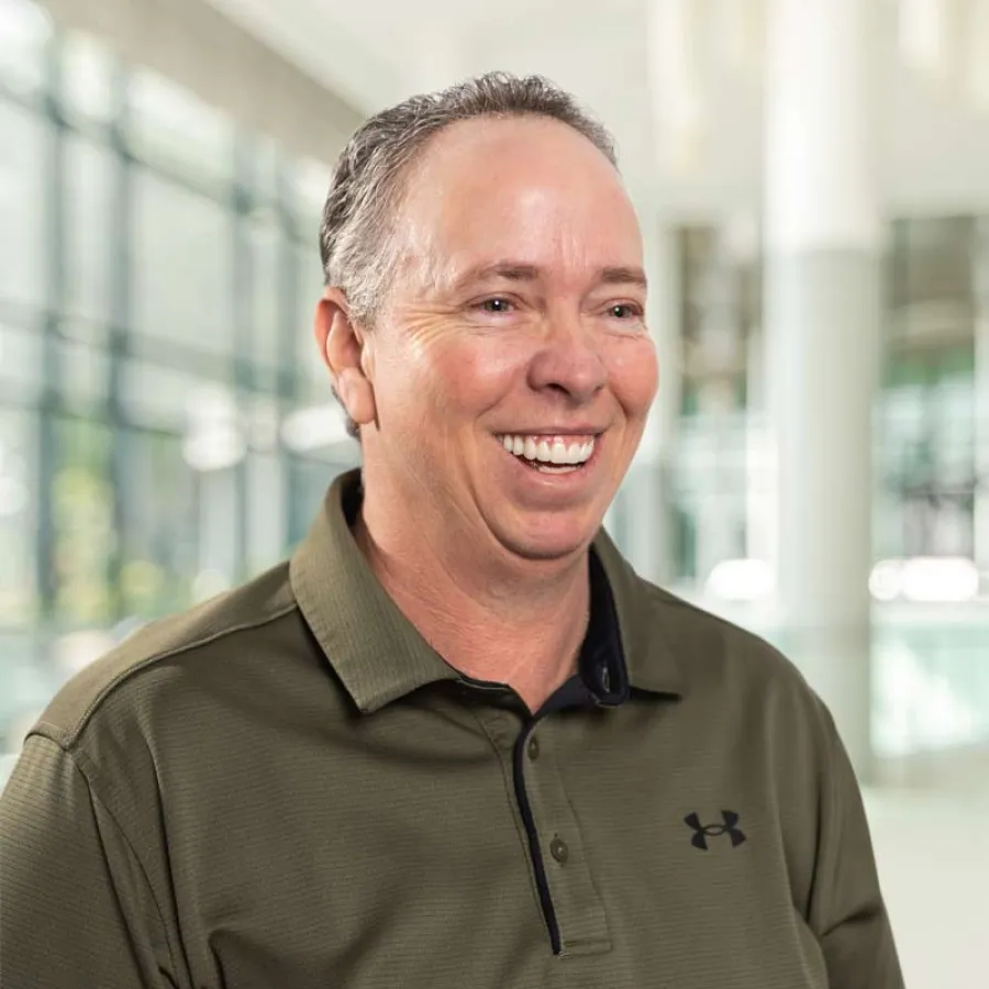 Middle-aged man in olive green Under Armour polo shirt smiling in bright modern indoor space.