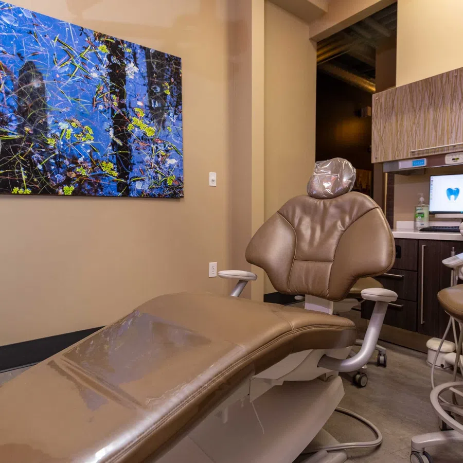 Modern dental exam room with brown chair, computer screen, artwork, and dental equipment in beige tones.