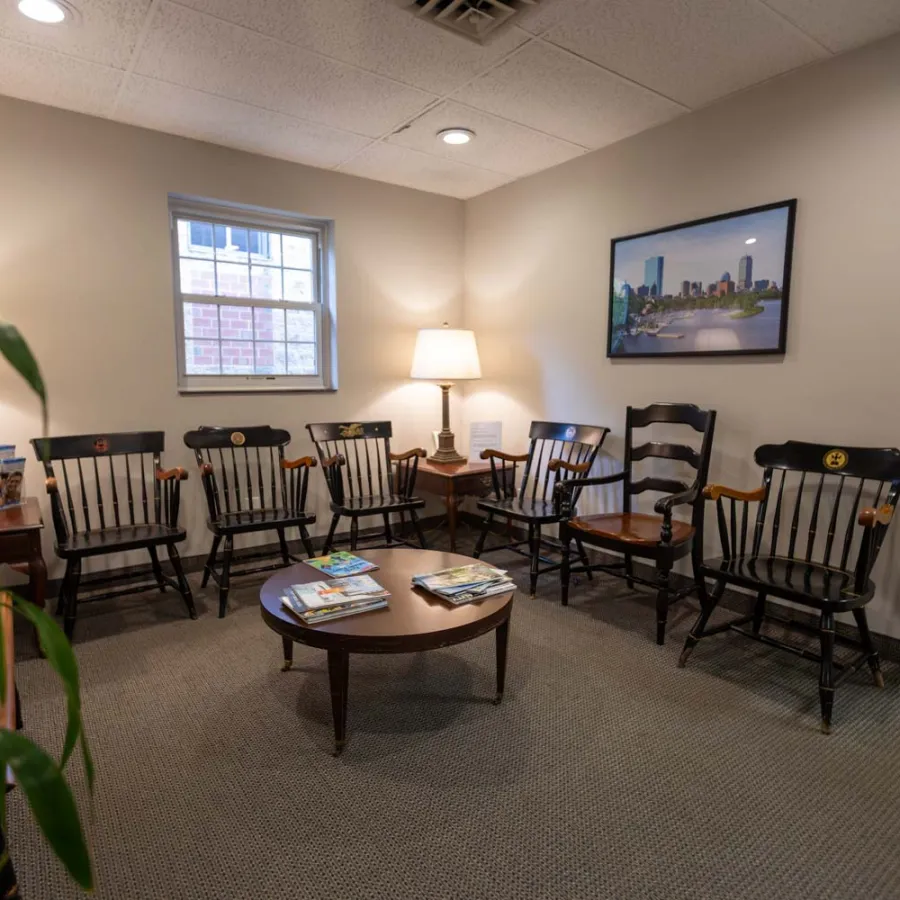 Waiting room with wooden chairs, a round table with magazines, lamps, and a cityscape framed picture on the wall.