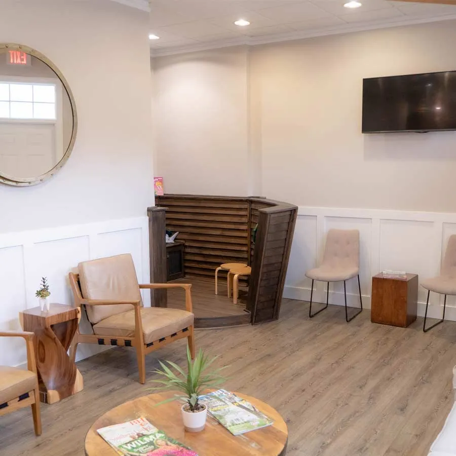 Modern waiting room with wooden chairs, a round table with magazines and plant, and a wall-mounted TV.