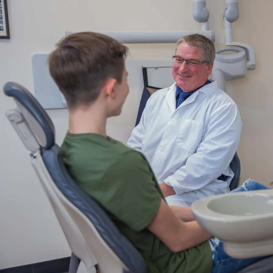 Dentist in white coat smiling while consulting a young male patient in a dental office.
