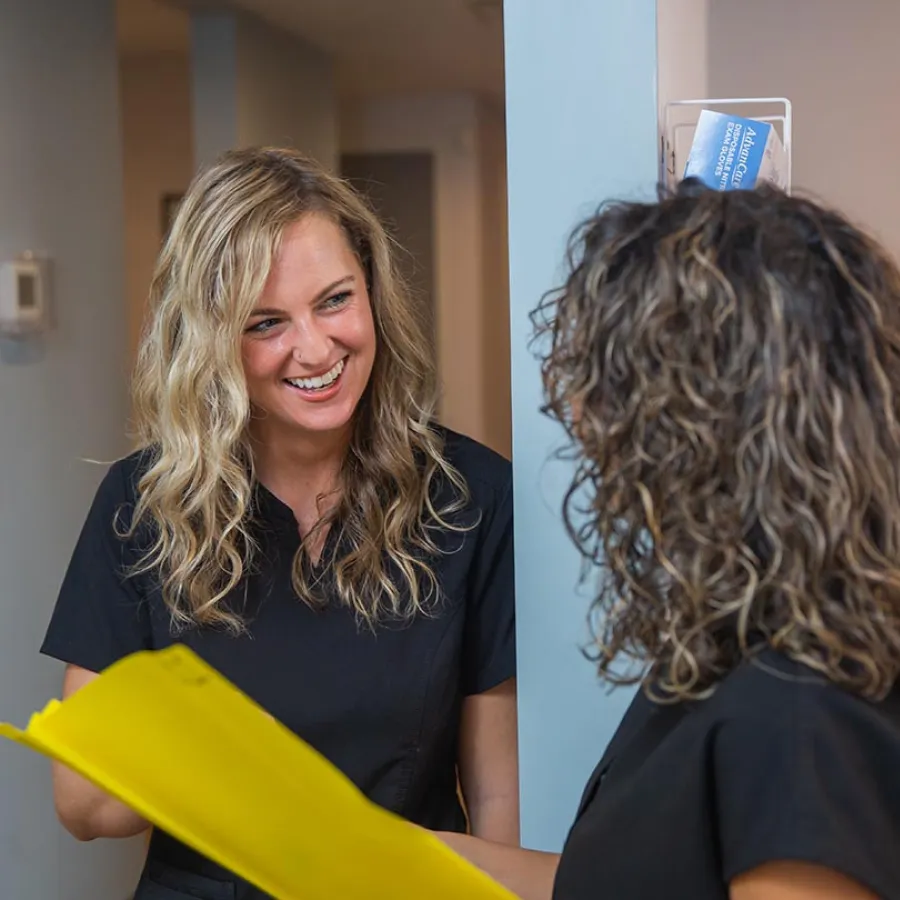 Two women in black scrubs review a yellow file folder while smiling and talking in a medical office.