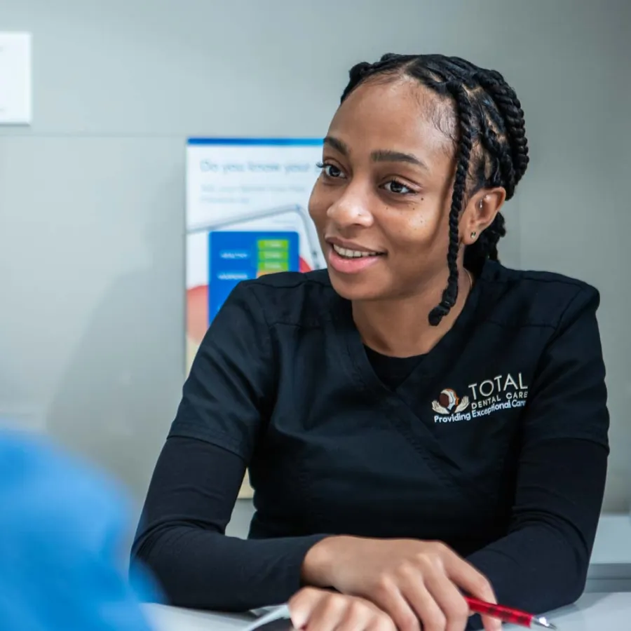 Smiling dental professional in black Total Dental Care uniform consulting a patient in a clinic setting.