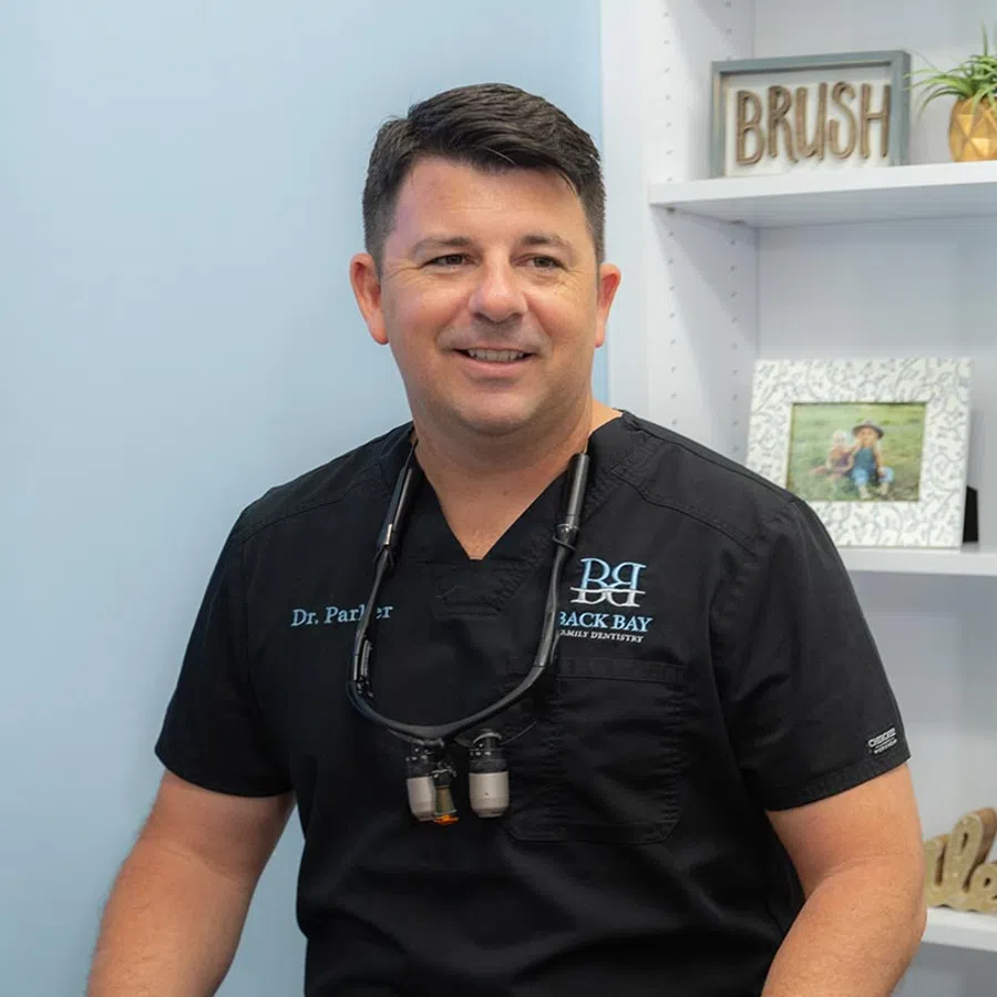 Smiling male dentist in black scrubs with magnifying glasses around neck in dental office setting.
