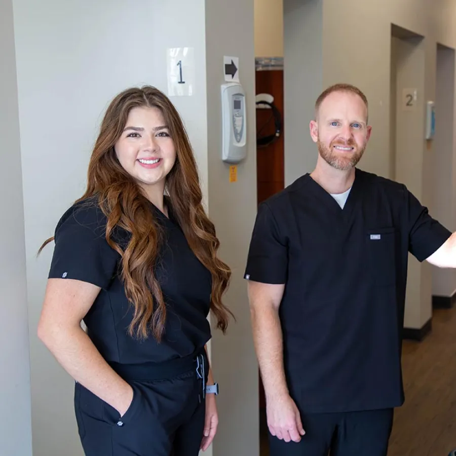 Two healthcare professionals in black scrubs smiling in a medical clinic hallway with numbered rooms.