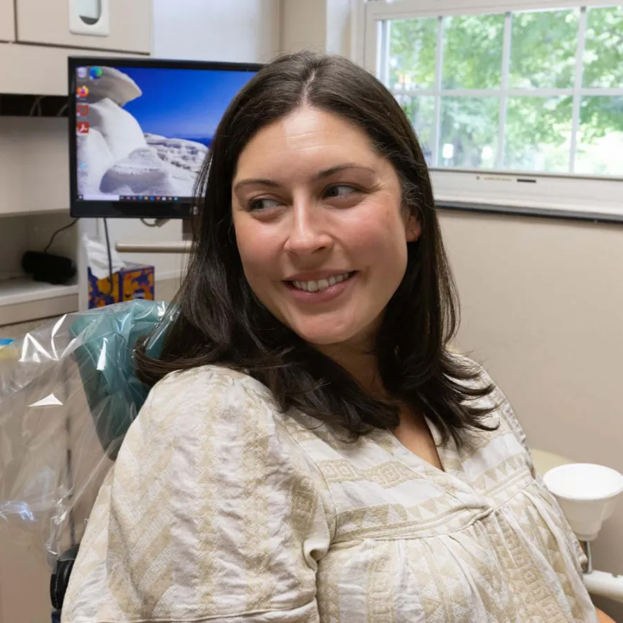 Smiling woman sitting in a dental chair in a bright dental office with computer and window in background