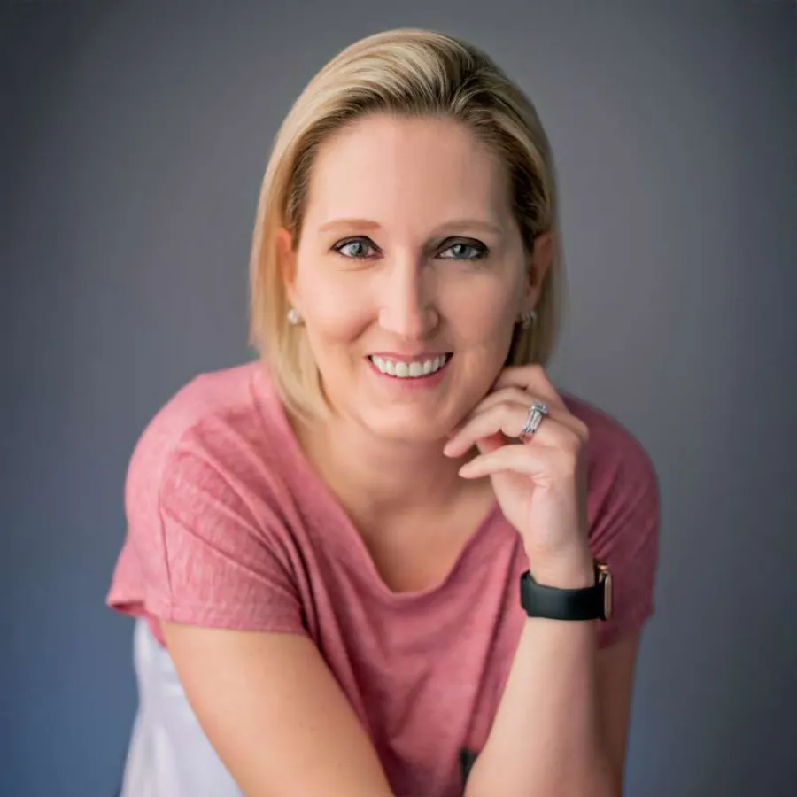 Smiling woman with short blonde hair wearing a pink top and smartwatch against a gray background.