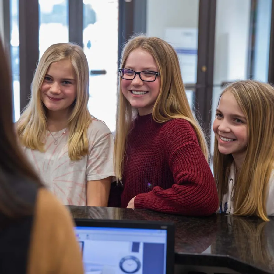 Three smiling teenage girls at a reception desk interacting with a staff member indoors in a bright setting.