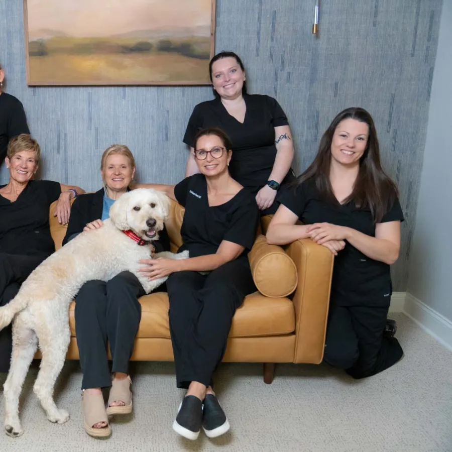 Group of seven women in black uniforms posing with a large white dog on a tan couch in a cozy room.