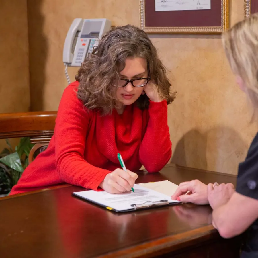Two women discuss and fill out paperwork at a desk in an office setting with framed certificates on the wall.