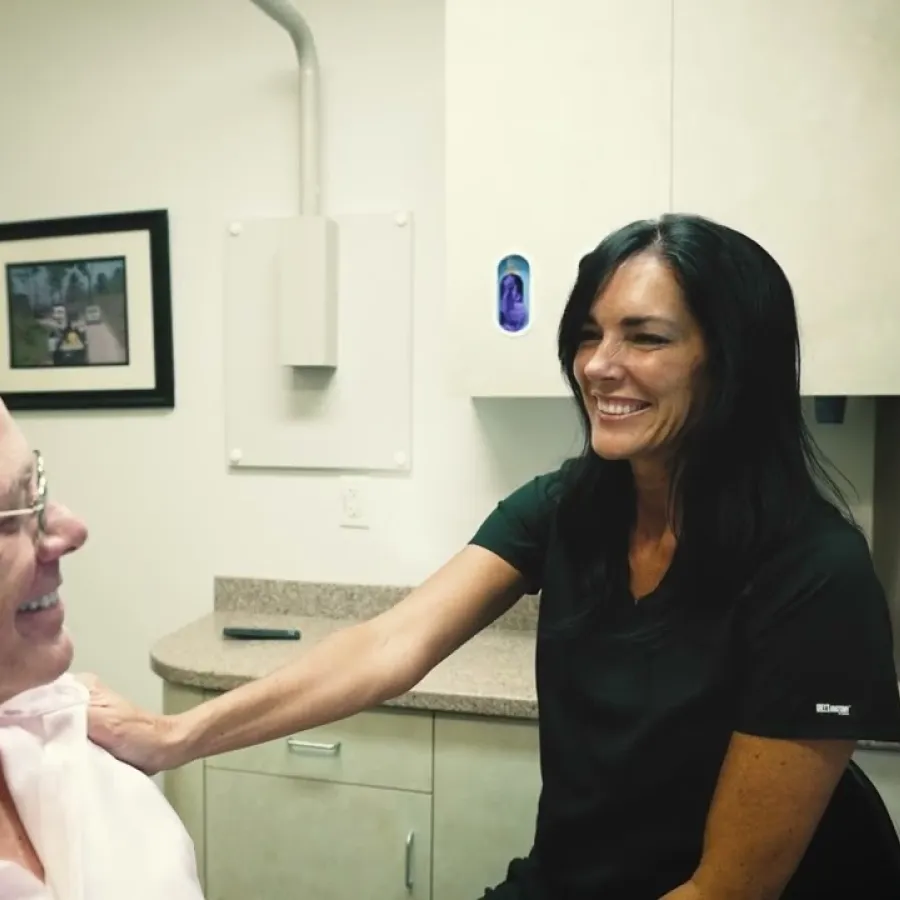 Smiling healthcare worker comforting elderly man in a medical office with framed pictures on the wall.