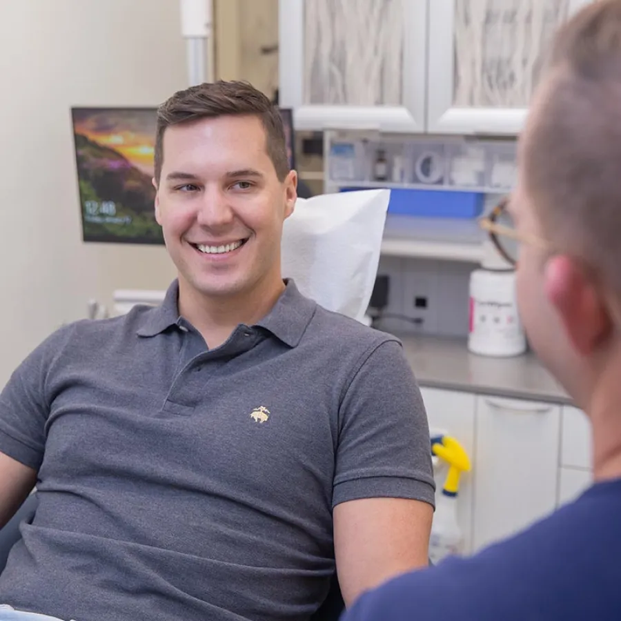 Smiling young man in a gray polo shirt sitting in a dental office chair talking to a dentist.