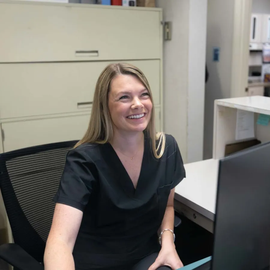 Smiling female healthcare worker in black scrubs sitting at a desk with computer in an office setting