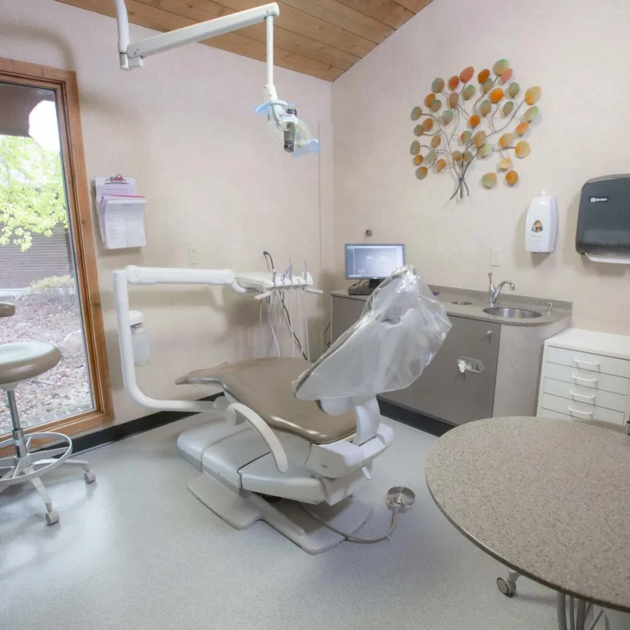 Modern dental office with patient chair, equipment, sink, computer, and large window with outdoor view.