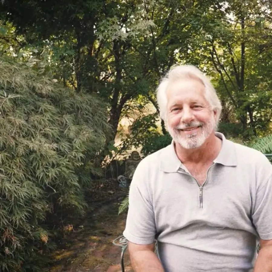 Smiling elderly man with white hair sitting on a metal chair in a lush green garden outdoors.