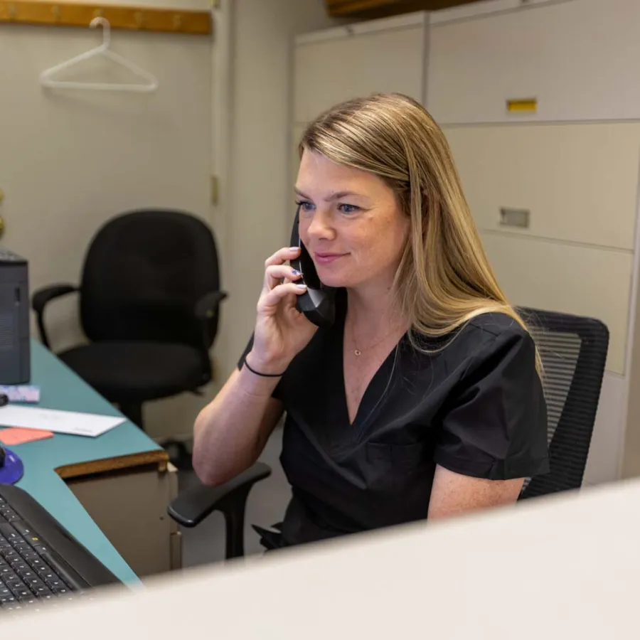 Woman in black scrubs sitting at desk using phone and computer in an office setting.